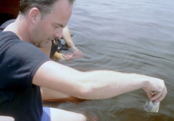 Lothar Frenz reicht vom Boot aus etwas ins Wasser, im Hintergrund schwimmt eine Person am Rio Negro