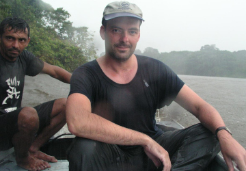 Lothar Frenz sitzt durchnässt im Boot bei Regen auf dem Amazonas, dahinter ein weiterer Insasse