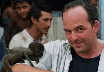 Lothar Frenz hält ein Wollaffenbaby auf dem Arm, im Hintergrund zwei Einheimische
