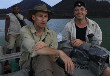 Lothar Frenz und ein Begleiter sitzen mit Filmausrüstung im Boot, im Hintergrund ein Vulkan